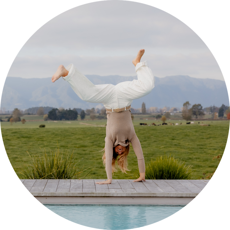 Vinka doing a cartwheel beside a pool, wearing white jeans, with rolling pasture and mountains behind.
